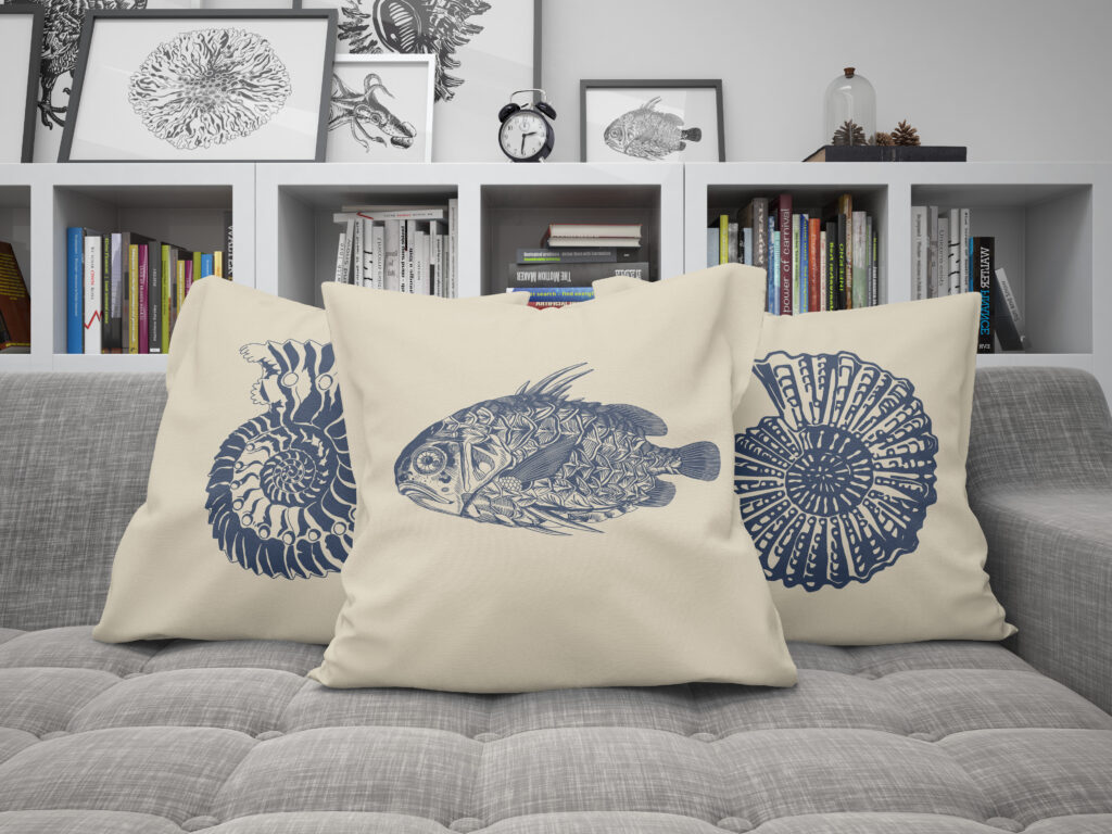 Drei beige Kissen, das in der Mitte ist mit einem Fisch bedruckt das links und rechts dahinter mit jeweils einem Ammoniten. Die Kissen liegen auf einem grauen Sofa. Im Hintergrund Regale mit Büchern und Bildern.