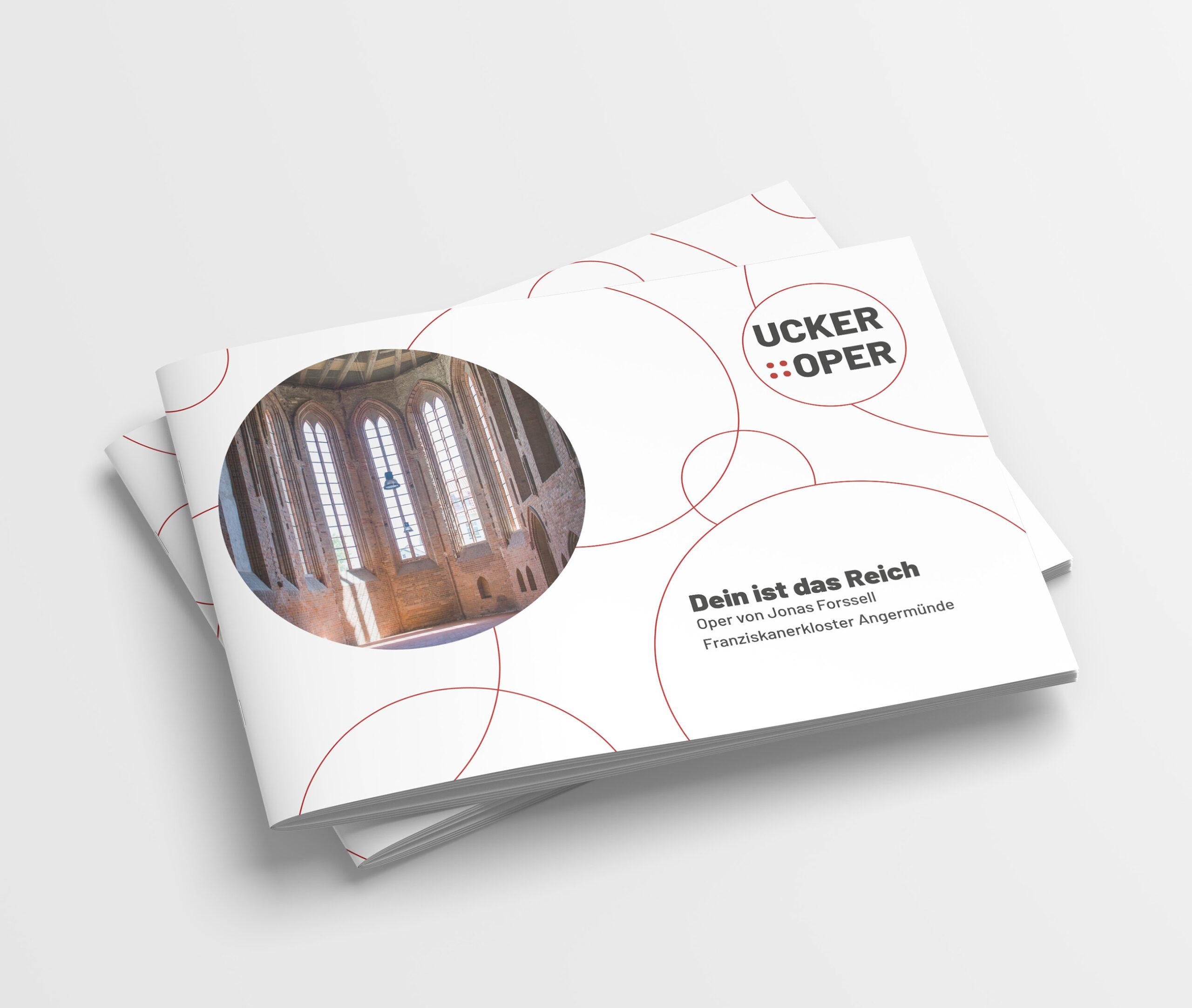 Mehrere Programmhefte der UckerOper auf einem Stapel. Das Cover ist mit Logo, roten Kreisen dem Stücktitel und einem Bild des Franziskanerklosters von Innen.